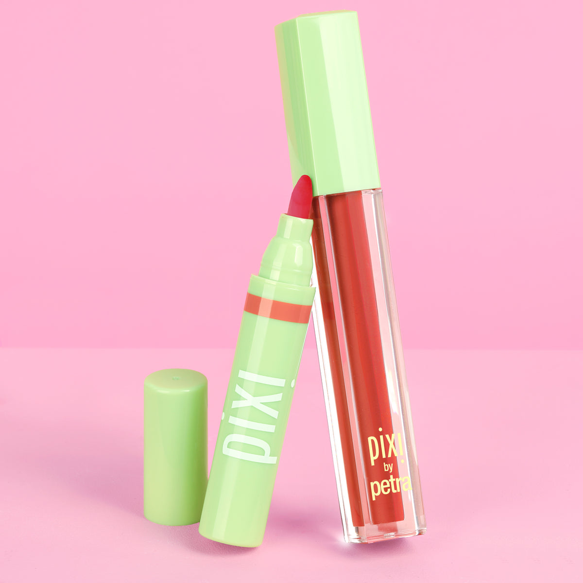 Pixi by Petra LipLift Max in a vibrant coral-red shade, shown in a clear tube with a light green cap next to its matching LipBlush against a solid pink background.