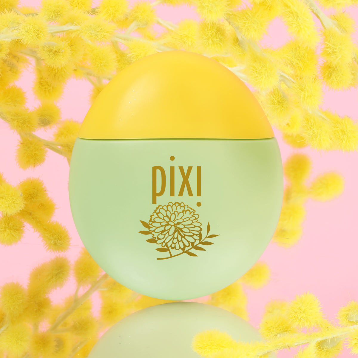 Pixi HandHero PixiMimosa scented hand lotion with mimosa blooms with a light green body and yellow cap, featuring a gold floral logo, set against a pink background with yellow flowers.