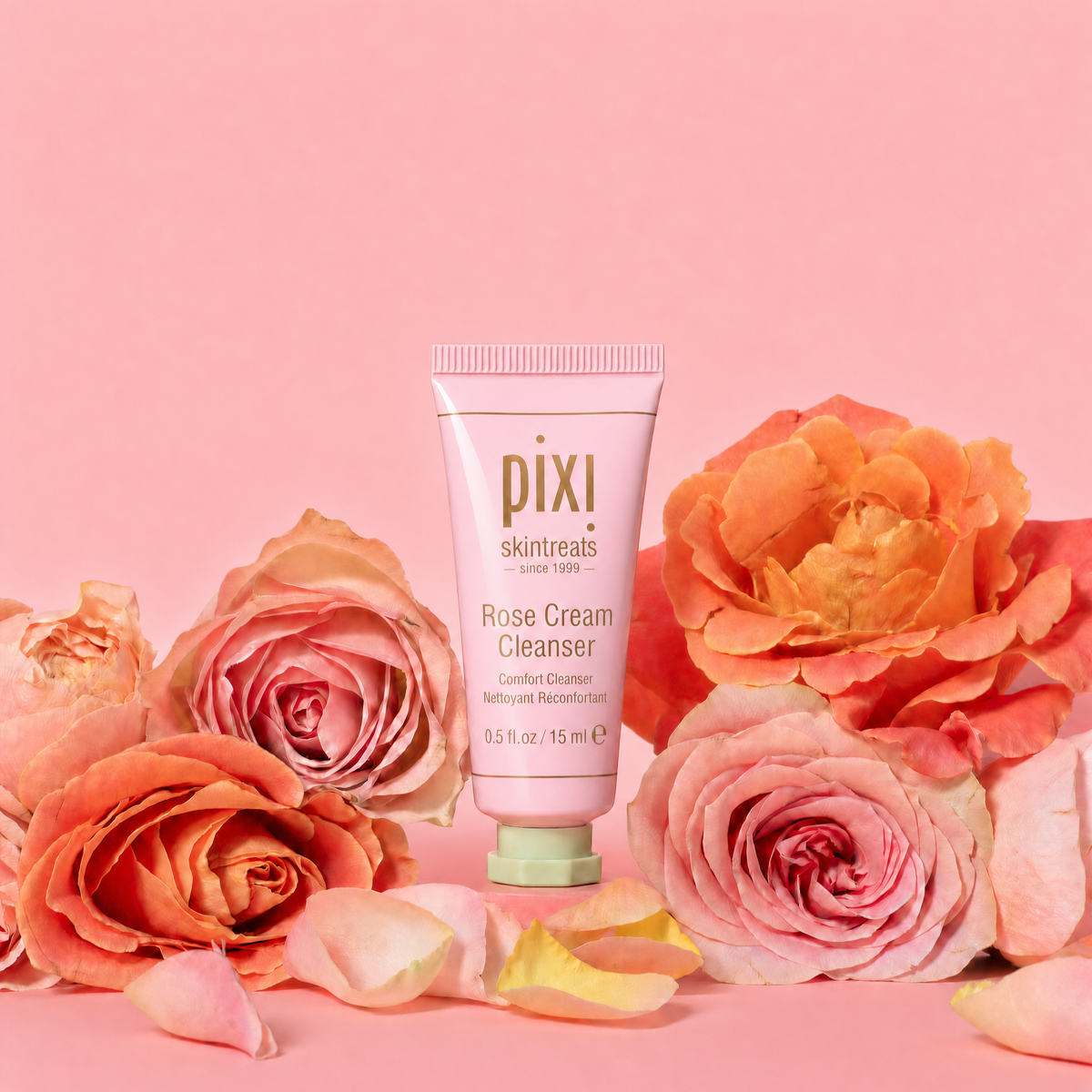 Pixi Skintreats Rose Cream Cleanser tube standing upright in the center, surrounded by large pink and peach roses with scattered petals on a soft pink background.
